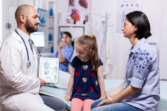 Pediatrician doctor explaining bones disease to mother using tablet with schelet image for sick child in hospital office. Physician specialist in pediatry medicine providing professional services