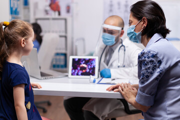 Fototapeta na wymiar Pediatrician wearing face mask and visor showing covid19 on tablet pc to mother during consultation. Practitioner in medicine providing professional radiographic treatment in hospital clinic