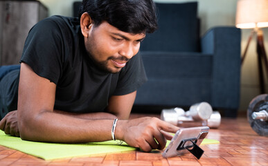 Fototapeta na wymiar Young man on yoga mat checking online workout or yoga tutorials on mobile phone at home - Concept of new normal, home gym due to coronavirus covid-19 pandemic lockdown.