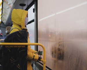 Fototapeta na wymiar A man in a yellow jacket on a bus with a hood on his head and a medical mask on his face looks out the misted window.