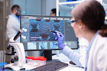 Professional biochemist analysing a medical experiment while working with her team to discover a vaccine. Healthcare physician pointing at virus on computer.