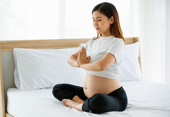 young pretty Asian woman sitting on a bed joins her hands in front of her chest with a smiley face...