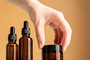 Set of unbranded cosmetics. Dark glass serum droppers and cream jar on a beige background. Female hand takes a jar with face cream. Beauty products.