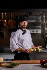 Handsome young male chef dressed in white uniform standing with finished cooked meal on plate,...