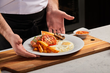 Breakfast on white plate in restaurant, copy space. fried eggs, roasted bacon, fries and vegetables prepared by professional chef cook. european cuisine concept. man putting plate on wooden board