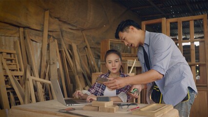 Asian carpenter or craftsman teach woman worker for wood working in workplace area and they look happy to discuss together. team are check quality the wood specification in product.