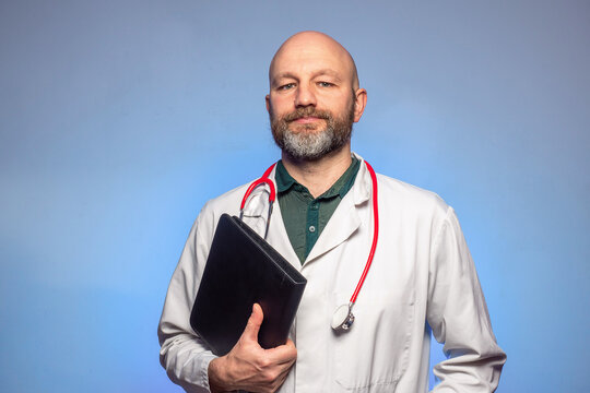 Bald Doctor With Beard On A Blue Background. The Model Is In His 40s. Looking At A Open Folder. Wearing White Uniform. Holding Black Folder.