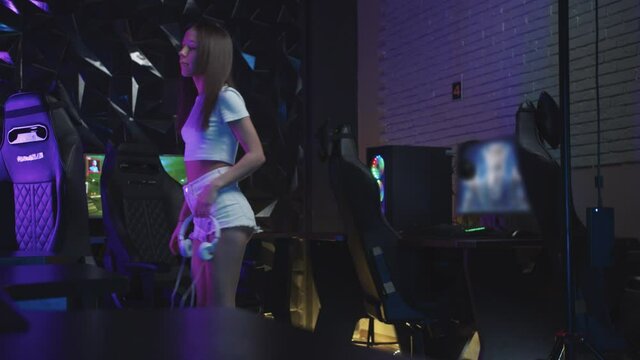 Girl gamer walks to the computer holding white headphones in the gaming club