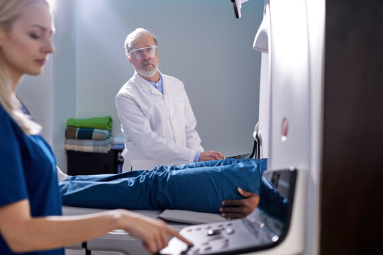 Doctor Assistant Turning On Complete CAT Scan System in Hospital Environment. Magnetic resonance imaging scan. Close-up hands and Equipment. focus on Senior Doctor in the Background