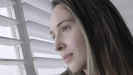 Close up of a woman looking out the window shutters. beautiful girl peeking out of blinds curious. spying on a neighbor and friend. lady hiding in the house behind blinds. morning light shines on face