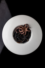 Black spaghetti with an octopus in the white plate, black background.