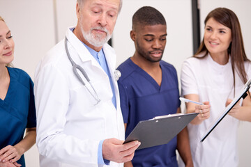 Obraz premium Senior doctor in white uniform consulting young doctors therapists using clipboard in hospital, Multidisciplinary medical team in white coats.