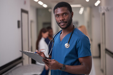 confident afro american nurse seriously posing in hospital hall aisle, take notes wearing blue...