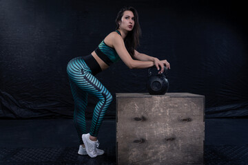 Fototapeta na wymiar a girl in sportswear poses against a dark background, leaning against an iron weight that stands on a wooden box