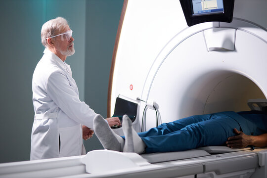 Senior Radiologist Controls MRI Or CT Or PET Scan With Male Patient Undergoing Procedure. High-Tech Modern Medical Equipment. Friendly Doctor Working With Patient