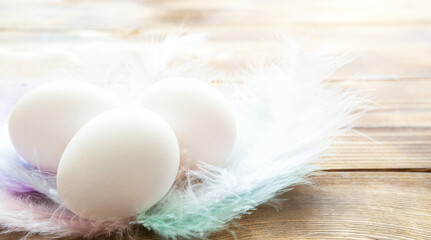 White chicken eggs on a wooden table in a nest of delicate pastel-colored feathers. Easter village background, natural eco-friendly products. Copy space