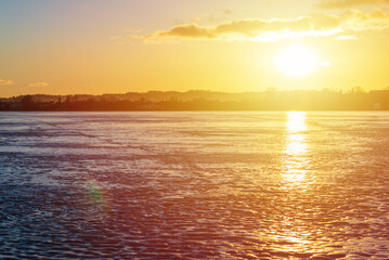 Winter landscape with frozen lake at sunrise or sunset. Lake glistening ice reflect a sun.Forest in the background.