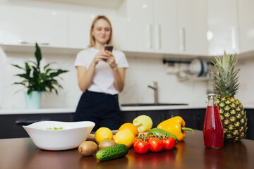 Young woman looking at new recipe in smartphone in kitchen. Girl cooking at home, preparing healthy vegetable salad. FOCUS ON FOOD.