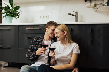 Happy young couple sitting on the floor in the kitchen and drinking red wine. They enjoying time together. 