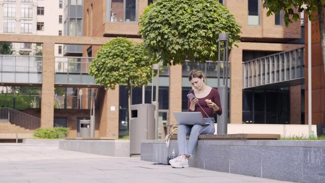 Young business woman with smartphone and modern laptop snacking in modern city park. At distance female on bench eating fast food eat while working remotely or studies online