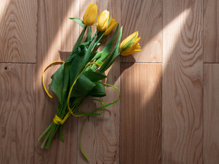 Beautiful yellow tulips on a wooden textured background, on a parquet. A bouquet of yellow flowers with green leaves on parquet boards in the rays of the spring sun. Spring yellow-green bouquet.