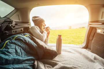 Girl resting in her car. Woman hiker, hiking backpacker traveler camper in sleeping bag, drinking hot tea and relaxing on top of mountain. Health care, authenticity, sense of balance and calmness.