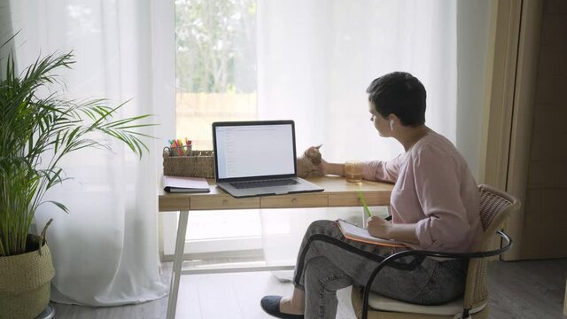 Woman with bluetooth earpiece writing in paper notebook at bamboo table with modern laptop against big window and petting cute Devon Rex cat relaxing on table