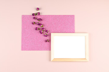 Wooden photo frame with white blank card and dry rosebuds on pastel pink background. Mock up poster frame. Stylish template
