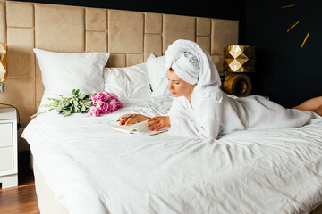 Woman in white bath robe and towel on her head lying on the bed and writing down important matters in a notebook.