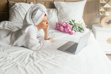 Obraz na płótnie Canvas Woman in white bath robe and towel on her head lying on the bed and watching movies on a laptop. Chill out and leisure concept.