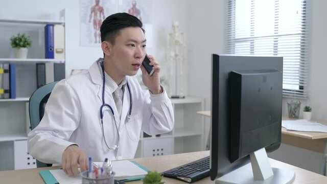 asian male doctor working in his office is taking a phone call and holding a pen on his chin while answering the question with concentration.