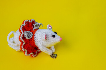 Knitted white rat in a red skirt on a yellow background. The rat holds a small copper heart in his hands. Cute handmade toy.