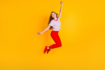 Fototapeta na wymiar Photo of pretty carefree lady jump raise arm wear striped shirt red pants footwear isolated yellow color background