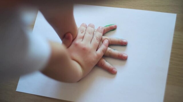 The hands of a little white-skinned boy leave colored spots of paint on white paper.  A white-skinned boy leaves traces of his hands on white paper.  European boy leaves traces of hands on white paper