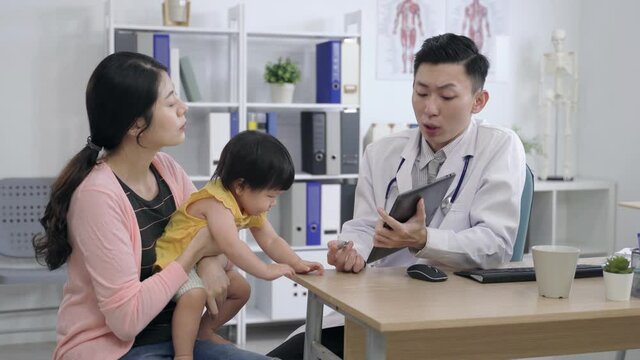 naughty asian baby girl is trying to take the pen away from the pediatrician as he is explaining the disease to the mother with a wireless pad at clinic.