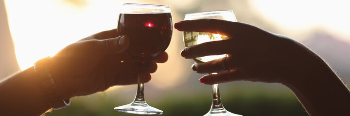 Male and female hands are holding glass of red and white wine against sunset background. Romantic...