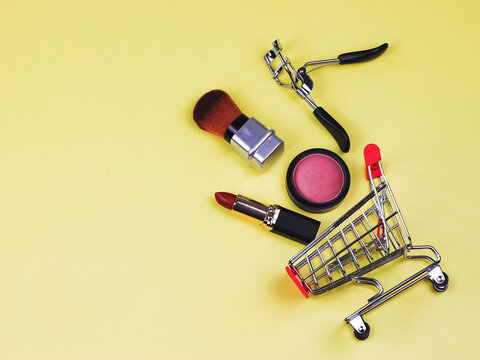  shopping trolley with makeup on yellow background. Blush, lipstick ,brush and eyelash curler in shopping cart. Cosmetic shopping concept.