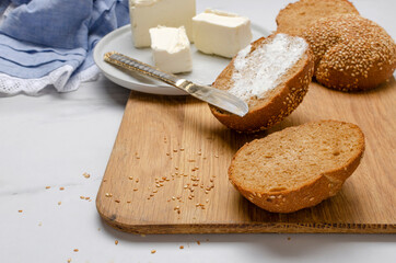 Closeup of rye buns, spread butter, knife and napkin on the wooden board on the white kitchen table
