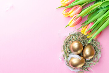Basket easter decoration: Golden eggs in basket with spring tulips, white feathers on pastel pink background. Congratulatory easter design. Top view.