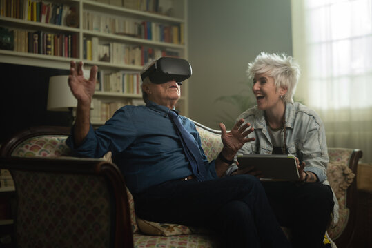 Cinematic shot of senior grandfather with vr glasses and granddaughter with tablet are having fun to use augmented reality technology together for family entertainment while sitting on sofa at home.