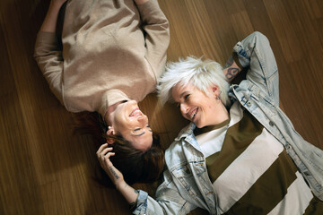 Cinematic shot of carefree happy homosexual female gay and transgender man couple in love is enjoying time together is speaking and caressing each other with affection while lying on floor at home.