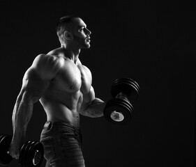 Fototapeta na wymiar Muscular well built men fitness trainer is standing sideways working out, lifting two dumbbells, does exercises for biceps looking forward copy space. Young man lifting weights. Black and white 