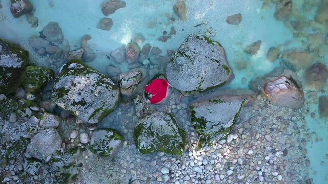 Young woman in red dress laying on a stone in the river. The drone floats quietly in the air, observing this piece of art. 