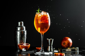 Aperol spritz cocktail in big wine glass with bloody oranges, red bitter, dry white wine, soda, zest and ice on dark background, bar tools