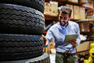 Bearded smiling supervisor standing in storage of shipping firm, holding tablet and checking on tires. Selective focus on tires. © Dusan Petkovic