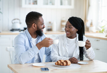 Cheerful young black spouses spending time in kitchen, having coffee with croissants