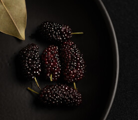Black mulberries on black dish with green leaf in frame