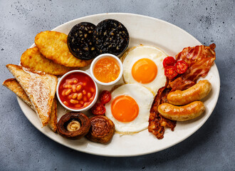 Full fry up English breakfast with fried eggs, sausages, bacon, black pudding, beans and toasts on gray concrete background
