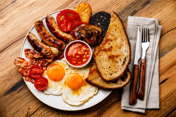 Full fry up English breakfast with fried eggs, sausages, bacon, black pudding, beans and toasts on wooden background