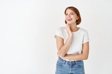 Beautiful candid girl with perfect smile, laughing and looking aside, touching natural beauty face without make-up, standing against white background